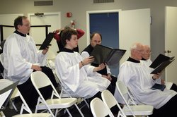 five men in white robes, seated, holding music and singing